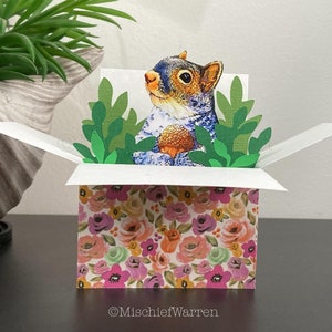 Könnte beinhalten: Eine Pop-up-Grußkarte mit einem Eichhörnchen, das eine Eichel hält, umgeben von grünem Papierlaub. Die Basis der Karte ist mit einem Blumenmuster aus rosa, orangefarbenen und violetten Blüten verziert. Der Text MischiefWarren steht unten.