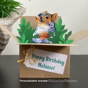 May include: A handmade birthday card with a pop-up squirrel holding an acorn, surrounded by green paper leaves. The brown card has a white tag with "Happy Birthday Melanie!" in green. A yellow bow is attached to the tag.