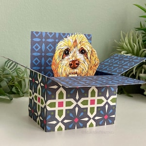May include: A pop-up greeting card with a golden doodle dog illustration emerging from a patterned box. The box is decorated with a geometric design in blue, white, green, and pink. The card is open, displaying the dog's face.