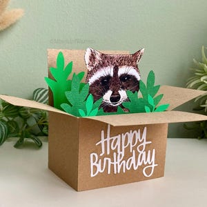 Pu&ograve; includere: Un biglietto di compleanno pop-up marrone con un procione che sbuca da dietro foglie di carta verdi. Il biglietto ha la forma di una scatola e presenta la scritta "Happy Birthday" in corsivo bianco.