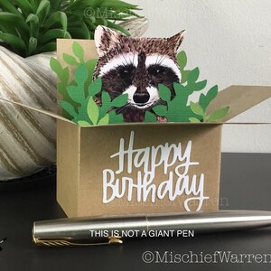 Pu&ograve; includere: Un biglietto di compleanno fatto a mano con un procione che sbuca da una scatola marrone con foglie verdi. Le parole "Happy Birthday" sono in scrittura bianca. Una penna d'argento con dettagli dorati &egrave; davanti al biglietto. Il testo "THIS IS NOT A GIANT PEN" &egrave; visibile.