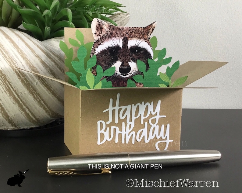 Pu&ograve; includere: Un biglietto di compleanno fatto a mano con un procione che sbuca da una scatola marrone con foglie verdi. Le parole "Happy Birthday" sono in scrittura bianca. Una penna d'argento con dettagli dorati &egrave; davanti al biglietto. Il testo "THIS IS NOT A GIANT PEN" &egrave; visibile.