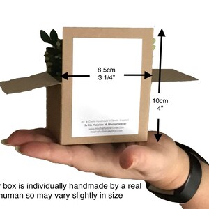 Puede incluir: Una peque&ntilde;a caja de cart&oacute;n abierta, sostenida en una mano, con dimensiones de 8,5 cm de ancho y 10 cm de alto. La caja contiene un peque&ntilde;o elemento decorativo verde y marr&oacute;n. El texto en la caja dice: "Cada caja est&aacute; hecha a mano individualmente por un ser humano real, por lo que puede variar ligeramente en tama&ntilde;o."