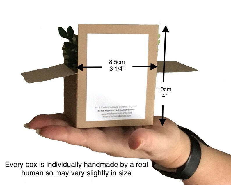 Puede incluir: Una peque&ntilde;a caja de cart&oacute;n abierta, sostenida en una mano, con dimensiones de 8,5 cm de ancho y 10 cm de alto. La caja contiene un peque&ntilde;o elemento decorativo verde y marr&oacute;n. El texto en la caja dice: "Cada caja est&aacute; hecha a mano individualmente por un ser humano real, por lo que puede variar ligeramente en tama&ntilde;o."