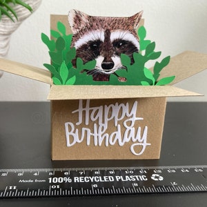 Pu&ograve; includere: Biglietto di compleanno fatto a mano con un procione che sbuca da una scatola marrone con foglie di carta verdi. Le parole "Happy Birthday" sono scritte in bianco sul fronte della scatola. Il biglietto &egrave; realizzato con materiali riciclati.