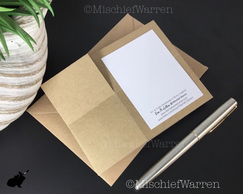 Pu&ograve; includere: Un biglietto di auguri con interno bianco &egrave; inserito in una busta di carta kraft. Una penna d'argento &egrave; appoggiata su una superficie nera accanto al biglietto. Il biglietto e la busta fanno parte di un set di cancelleria. Il testo "Mischief Warren" &egrave; visibile.