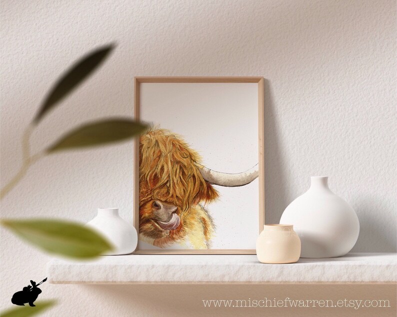 Puede incluir: Una pintura de acuarela de una vaca Highland con pelo largo y marr&oacute;n y peludo. La vaca est&aacute; mirando hacia un lado con la lengua fuera. La pintura est&aacute; enmarcada en un marco de madera simple.