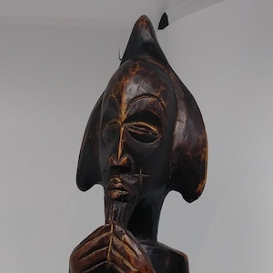 Puede incluir: Una escultura de madera marrón oscuro de una persona con la cabeza puntiaguda y una barba larga y fina. La escultura está de pie con las manos juntas frente al pecho.