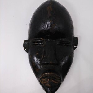 May include: A black wooden mask with a carved face. The mask has a long, narrow shape and features a prominent nose, closed eyes, and a wide mouth.