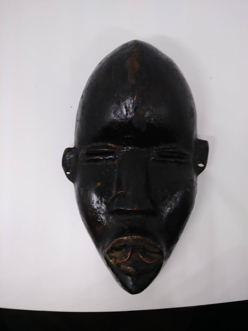 May include: A black wooden mask with a carved face. The mask has a long, narrow shape and features a prominent nose, closed eyes, and a wide mouth.