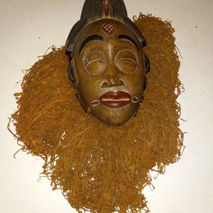 Op de afbeelding: Een houten masker met gesloten ogen, rode lippen en een bruine teint. Het masker heeft een donkere hoofdbedekking met een rode streep en een decoratief element op het voorhoofd. Het is omgeven door een textuurachtig, stro-achtig materiaal.