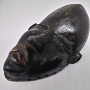 May include: A dark brown wooden mask with a carved face. The mask has a long, narrow shape and is slightly curved. The eyes are closed and the mouth is slightly open. The mask has a series of small holes around the edges.