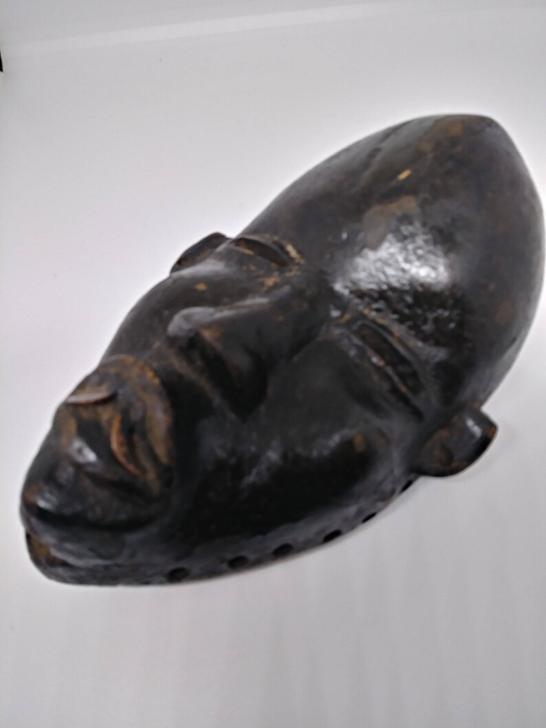 May include: A dark brown wooden mask with a carved face. The mask has a long, narrow shape and is slightly curved. The eyes are closed and the mouth is slightly open. The mask has a series of small holes around the edges.