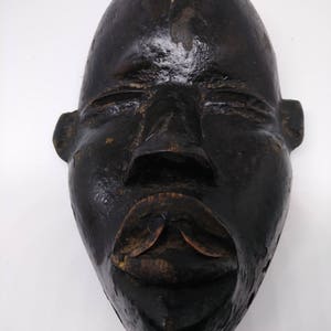 May include: A dark wooden mask with a carved human face. The mask has a wide, flat forehead, closed eyes, and a large, open mouth with a carved lip.