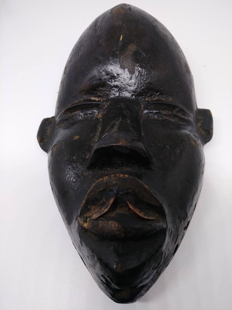 May include: A dark wooden mask with a carved human face. The mask has a wide, flat forehead, closed eyes, and a large, open mouth with a carved lip.