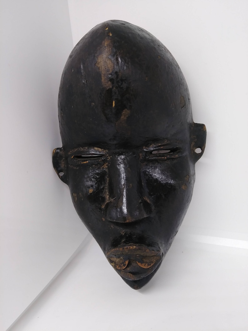 May include: A black wooden mask with a human face. The mask has a long, thin nose and a wide mouth with a carved lip. The eyes are closed and the mask has a smooth, polished surface.