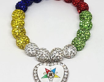 May include: A colorful beaded bracelet with a heart-shaped pendant. The bracelet features a rainbow of colors, including yellow, blue, red, and green, with a silver-tone heart pendant adorned with a colorful emblem and clear rhinestones.