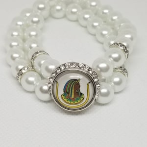 May include: A white pearl bracelet with a silver charm featuring a colorful image of an Egyptian queen. The charm is surrounded by rhinestones.