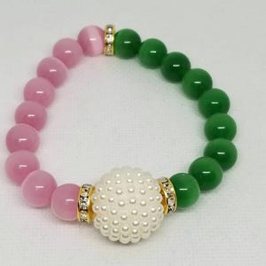 May include: A beaded bracelet featuring pink and green beads, with a central white pearl-studded bead. Gold-colored spacers with clear crystals accent the design. The bracelet is a stylish accessory.