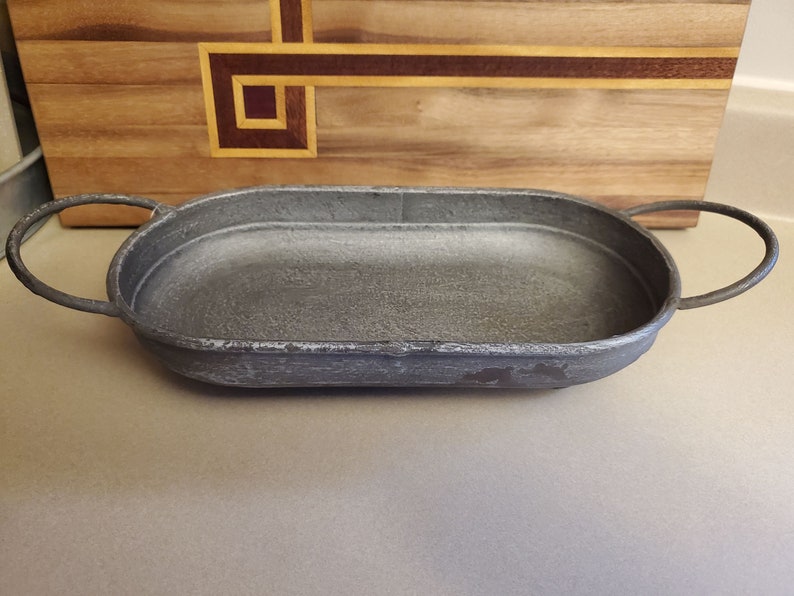 Pode incluir: Uma bandeja oval de metal cinza com duas al&ccedil;as. A bandeja tem um acabamento desgastado e &eacute; perfeita para servir petiscos ou bebidas.