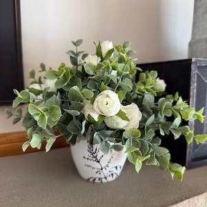 May include: A white ceramic mug with a black handle and the text "But First, Coffee" in a black, bold font. The mug is on a white surface.