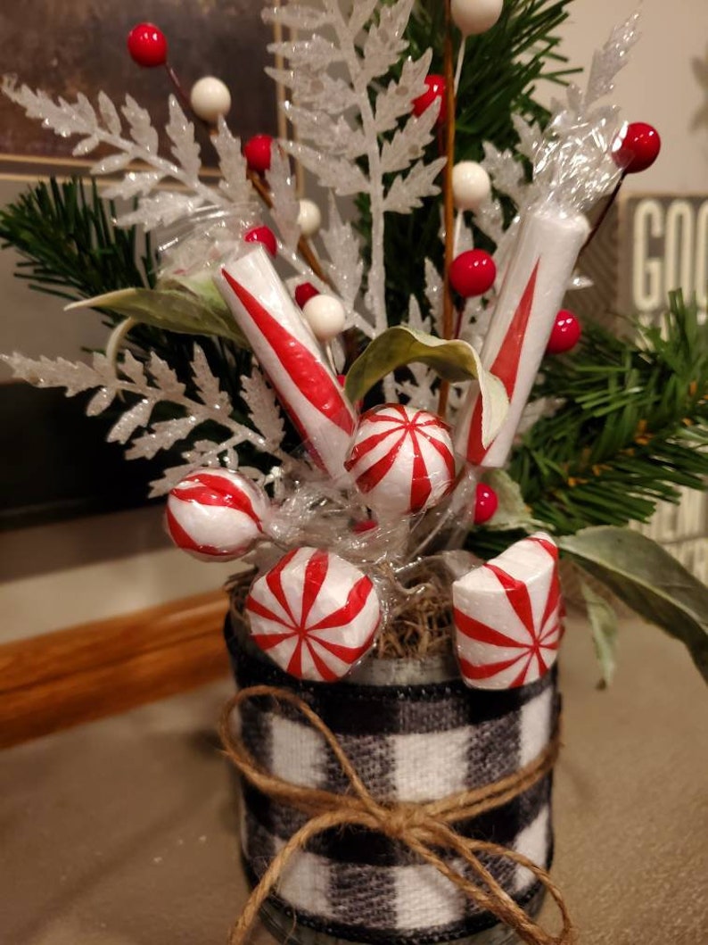 Pode incluir: Um pequeno frasco de vidro decorado com uma fita xadrez preta e branca e barbante, cheio de vegeta&ccedil;&atilde;o artificial, bagas vermelhas e doces de menta branca e vermelha.