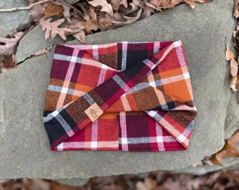May include: A folded plaid bandana in shades of red, orange, black, and white with a brown leather tag. The bandana is folded into a triangular shape and has a white border.