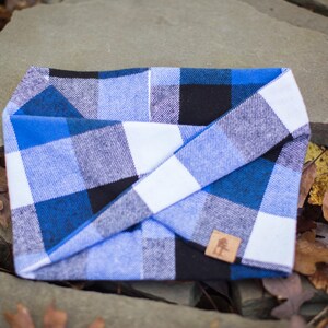 May include: A blue, black, and white plaid dog bandana with a brown leather tag featuring a tree logo.