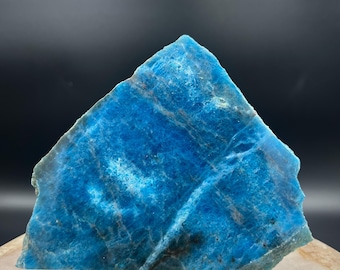 May include: A vibrant blue apatite gemstone slab with a rough, natural texture. The stone is a striking shade of turquoise, with lighter blue and white veining. The slab is displayed on a clear stand, showcasing its unique crystalline structure.