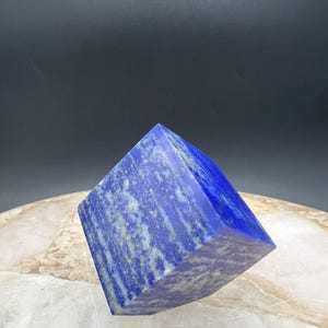 May include: A faceted, blue lapis lazuli crystal. The gemstone has a deep blue color with streaks of white and gold. The crystal is cut into a square shape with a pointed top. The background is a neutral gray.
