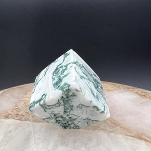 May include: A white and green moss agate crystal pyramid. The stone features a mix of white and translucent areas with green, moss-like inclusions. The pyramid shape has a flat base and a pointed top, showcasing the natural patterns.