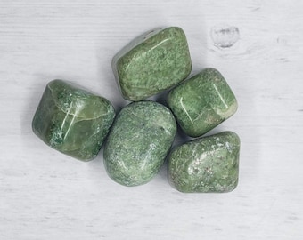 May include: Five polished green stones with varying shapes and sizes. The stones have a mottled appearance with lighter and darker green areas. The stones are arranged on a light-colored wooden surface.