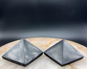 May include: Two dark gray pyramid-shaped objects are displayed on a light-colored, patterned surface. The pyramids have a square base and a pointed top. The background is a dark, solid color.