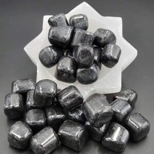 May include: A collection of polished, black tourmaline stones. The stones are tumbled and smooth, with a glossy sheen. They are displayed in a white, star-shaped bowl, with additional stones scattered around the base. The background is a solid black.