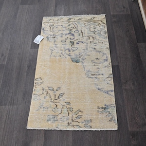 May include: A rectangular, vintage-style rug with a beige background and intricate blue and brown patterns. The rug has a distressed look, with visible wear and tear, and is placed on a dark wooden floor. A tag is attached to the rug.