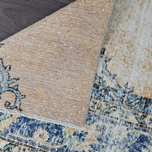 Op de afbeelding: Een close-up van de achterkant van een tapijt met een beige achtergrond en een blauwe en witte gepatroneerde rand. Het tapijt is dubbelgevouwen, waardoor de beige achterkant zichtbaar is.