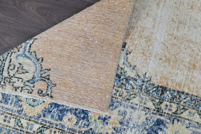 Op de afbeelding: Een close-up van de achterkant van een tapijt met een beige achtergrond en een blauwe en witte gepatroneerde rand. Het tapijt is dubbelgevouwen, waardoor de beige achterkant zichtbaar is.