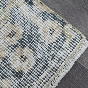 May include: Close-up of a woven rug with a fringed border. The rug features a geometric pattern in shades of beige, blue, and white.