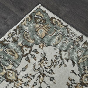Op de afbeelding: Een vintage tapijt met een cr&egrave;mekleurige achtergrond en een rand van ingewikkelde bloemmotieven in tinten groen, bruin en beige.
