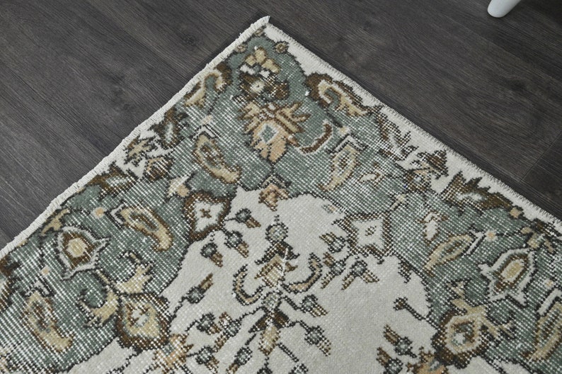 Op de afbeelding: Een vintage tapijt met een cr&egrave;mekleurige achtergrond en een rand van ingewikkelde bloemmotieven in tinten groen, bruin en beige.