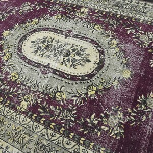 May include: A vintage rug with a faded floral pattern in shades of purple, cream, and black. The rug has a worn and distressed look, with some areas showing signs of wear and tear.