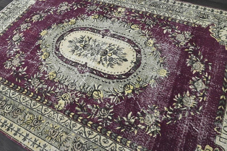 May include: A vintage rug with a faded floral pattern in shades of purple, cream, and black. The rug has a worn and distressed look, with some areas showing signs of wear and tear.