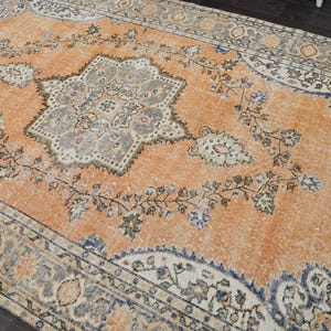 Puede incluir: Una alfombra rectangular antigua con un aspecto desgastado y descolorido. La alfombra presenta un medall&oacute;n central en gris y crema, rodeado por un campo naranja con motivos florales y de vid. El borde es una mezcla de azul, crema y gris.