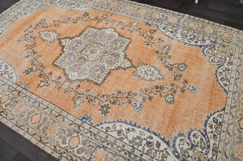 Puede incluir: Una alfombra rectangular antigua con un aspecto desgastado y descolorido. La alfombra presenta un medall&oacute;n central en gris y crema, rodeado por un campo naranja con motivos florales y de vid. El borde es una mezcla de azul, crema y gris.