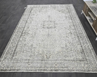 Puede incluir: Una alfombra rectangular grande con un patrón desgastado y descolorido en tonos crema, gris claro y carbón. El diseño incluye un medallón central rodeado de intrincados motivos florales y de enredaderas con un borde decorativo ancho.