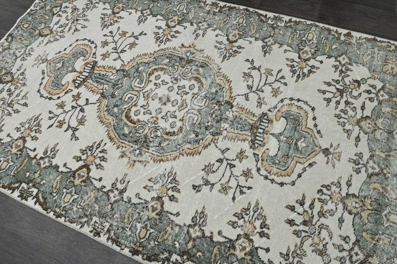 Op de afbeelding: Een vintage tapijt met een cr&egrave;mekleurige achtergrond en een gedetailleerd bloemenpatroon in tinten groen en bruin.