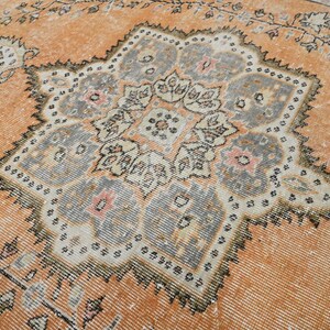 Puede incluir: Una alfombra antigua y desgastada con un dise&ntilde;o de medall&oacute;n central. La alfombra presenta un fondo naranja c&aacute;lido con un patr&oacute;n central detallado de varias capas en tonos grises, crema y rosa. Los bordes de la alfombra est&aacute;n adornados con motivos florales.