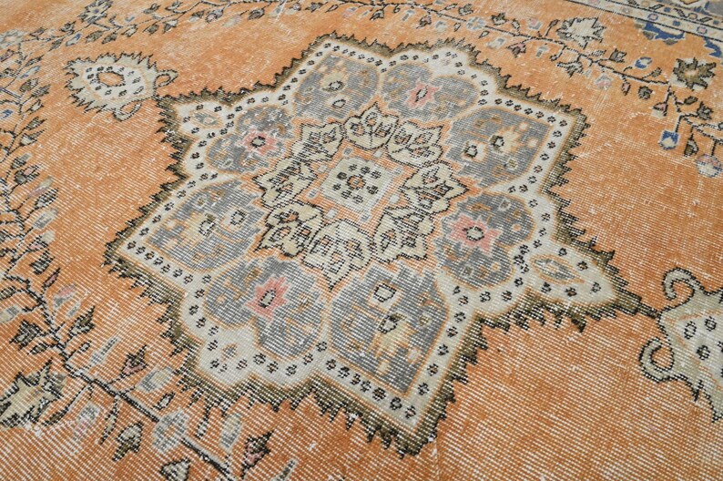 Puede incluir: Una alfombra antigua y desgastada con un dise&ntilde;o de medall&oacute;n central. La alfombra presenta un fondo naranja c&aacute;lido con un patr&oacute;n central detallado de varias capas en tonos grises, crema y rosa. Los bordes de la alfombra est&aacute;n adornados con motivos florales.