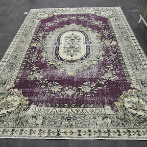 May include: A vintage-style rug with a faded purple background and intricate floral patterns in cream and beige. The rug has a distressed look and a worn border.