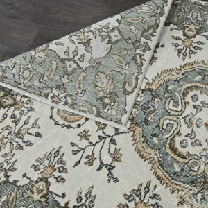 Op de afbeelding: Een vintage tapijt met een witte achtergrond en een groen, bruin en beige bloemenpatroon.